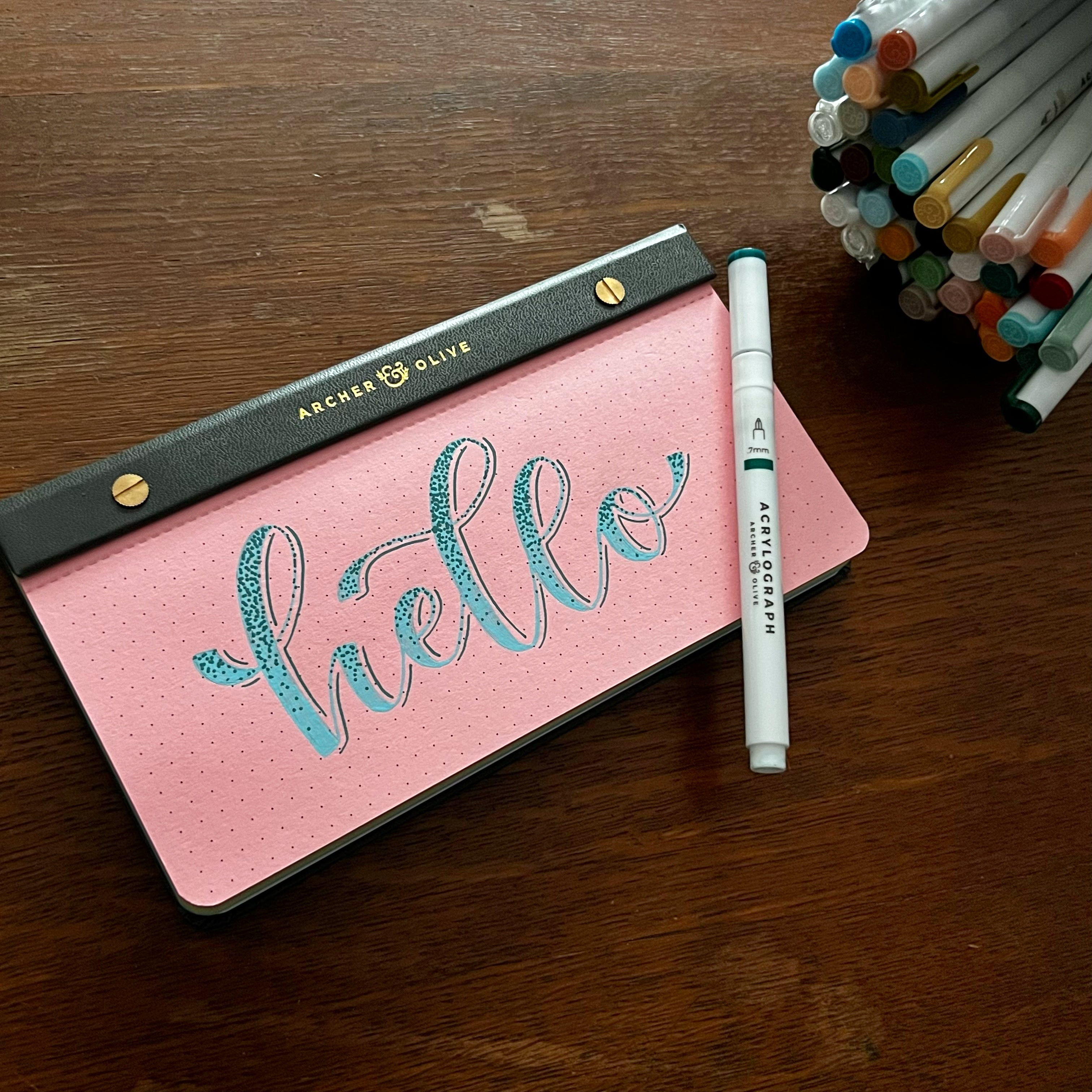 Pink paper notepad with container of Acrylograph pens. Faux calligraphy “hello” lettered on the paper.